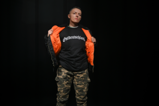Person auf Bühne öffnet Jacke und zeigt T‑Shirt mit Aufschrift „Ostdeutschland“, trägt Tarnhose, dunkler Hintergrund.