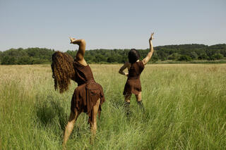 Das Bild bildet die beiden Künstlerinnen tanzend in einem Feld ab. Beide tragen braune Kleider, die Arme tänzerisch nach oben gerichtet, den Rücken zur Kamera. Die Umgebung ist nur Kornfeld, am Horizont Bäume und blauer Himmel. 