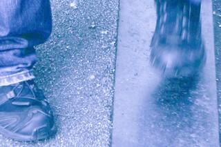 Auf dem Bild sind schwarze Schuhe und eine Jeanshose zu sehen. Sie stehen auf Asphalt und Metall. Das Bild ist blau-violett eingefärbt.