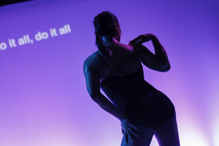 Ein*e Tänzer*in in einem schwarzen Body steht vor einem lila beleuchteten Hintergrund, auf dem der Text „do it all, do it all“ in Weiß zu sehen ist.
