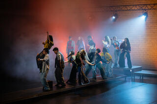 Eine Gruppe junger Tänzer in lässiger Alltagskleidung performt auf einer Bühne vor einer Ziegelwand mit atmosphärischem Licht und dichtem Bühnennebel.